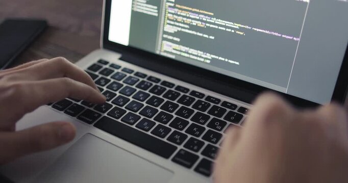 Close-up of hands typing on a laptop with blurred programming code on the screen and a smartphone lying beside the computer.