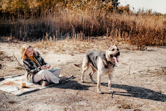 Smiling woman holding sketchbook and looking at dog outdoors. Happiness and strong bond with domestic animal in nature.