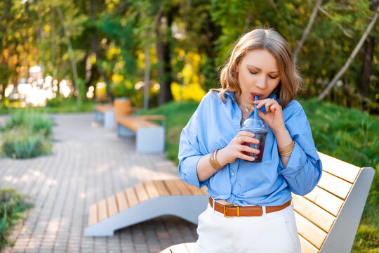 Woman walking in sunny park drinking iced beverage from plastic cup with straw. Refreshing cold drink and casual summer walk.