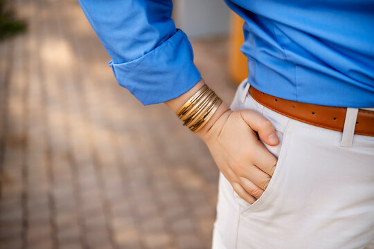 Woman hand in pocket of white pants with gold bracelets and blue shirt outdoors. Casual summer fashion and stylish accessories.