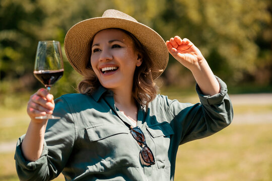 Happy smiling woman in straw hat holding glass of red wine outdoors in sunny park. Cheerful mood and joyful summer picnic vibe.