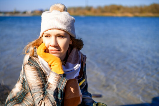 Thoughtful woman sitting by lake in autumn in beanie and plaid shirt resting head on hand. Melancholy mood and quiet solitude in nature.