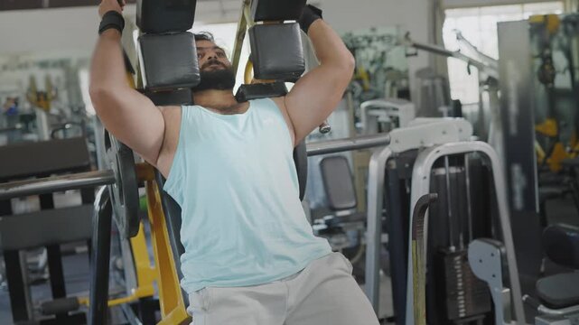 A young Indian Asian fit muscular male performs a leg squat exercise using a machine as part of resistance or weight training in a modern gym. Fitness, workout, healthy lifestyle, wellness concept