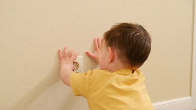 Child boy touching electric socket on wall. Dangerous behavior at home. Domestic toddler safety concern. Risk of electrical shock in house environment. Need for home childproofing.
