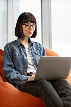 A woman with glasses and a bob haircut sits comfortably on an orange beanbag chair, typing on her laptop in a well-lit room