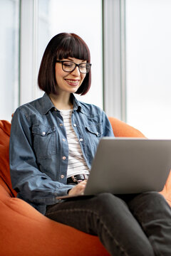 A young woman with glasses and a bob haircut smiles while working on her laptop computer, seated comfortably on an orange beanbag chair