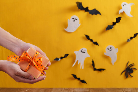 Cropped hands holding Halloween gift box on yellow background with paper ghosts and bats