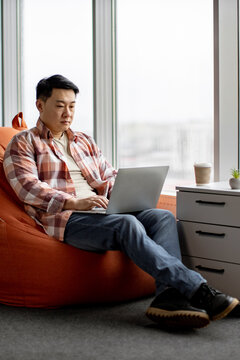 A man sits comfortably in a beanbag chair near a large window, typing on his laptop and looking focused on his work