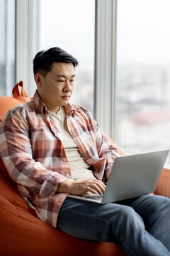 A man sits comfortably in an orange beanbag chair, focused on his laptop screen while looking out a large window