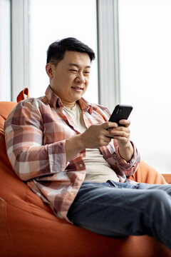 A man sits comfortably on an orange beanbag chair while looking at his smartphone, with natural light streaming in from a window