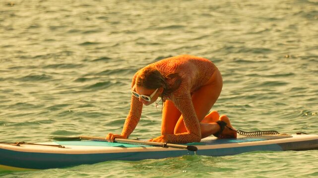 Paddleboarding woman kneeling on inflatable SUP board during golden hour enjoying summer ocean vacation