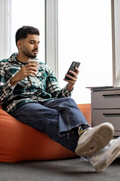 A young man in a plaid shirt sits comfortably on an orange beanbag chair, holding a coffee cup and looking at his smartphone