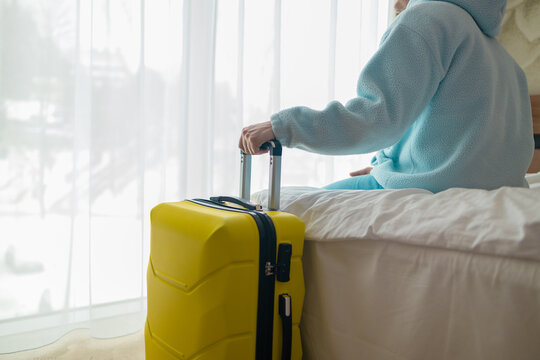 Side view of a male traveler holding luggage handle while resting on a couch or bed near window. Concept of travel arrival, hotel check-in, cozy tourism accommodation, and departure waiting.