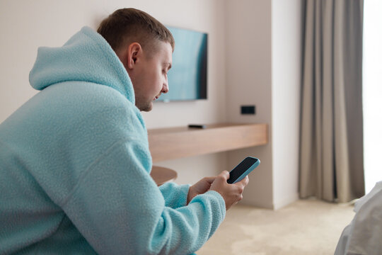 Side view profile of a millennial male traveler checking mobile phone screen in cozy bedroom. Concept of digital booking, searching information, online navigation, and remote work.