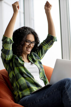 A young woman with curly hair and glasses smiles broadly while raising her arms in triumph as she looks at her laptop screen