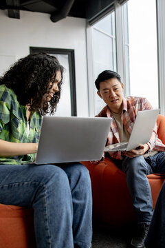 Two young adults, a woman and a man, are sitting on beanbag chairs and actively working together on their laptops in a bright, modern space