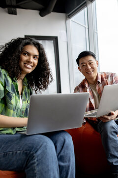 A young woman and a man of Asian descent smiling while working together on laptops in a bright, modern office space