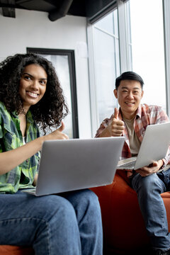 A woman and a man of diverse backgrounds smiling and giving thumbs up while working on laptops together indoors