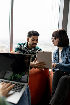 Two professionals intently discussing a project on a tablet while another person works on a laptop in a modern office setting
