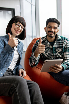 A man and woman in casual attire sit comfortably on beanbag chairs, smiling and giving a thumbs-up gesture while holding a tablet