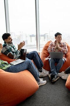 A diverse group of professionals sits on beanbag chairs near a large window, clapping and smiling during a meeting