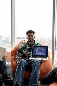 A young man in a plaid shirt presents a car blueprint on a laptop while sitting in a beanbag chair near a large window