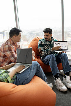 Diverse colleagues engaged in a discussion while working on laptops in a modern office setting with large windows