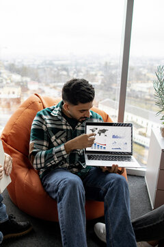 A man in a plaid shirt points to a laptop screen displaying world map and financial charts with a downward trend