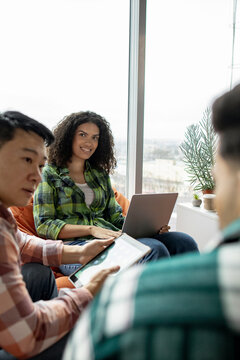 A group of young professionals in casual attire are gathered around a laptop and tablet, discussing ideas near a large window
