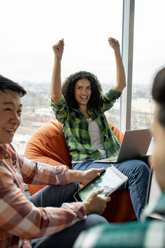 A diverse group of young adults expresses joy and excitement while working together on a project, using technology indoors