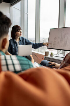 A woman in glasses points to a building blueprint on a large monitor while presenting to colleagues in a modern office setting