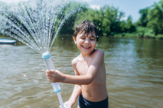 Happy boy sprays water with a toy pump in the river during summer