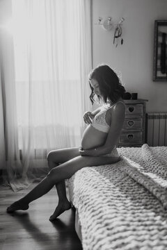Pregnant woman sits on the bed and touches her baby bump indoors