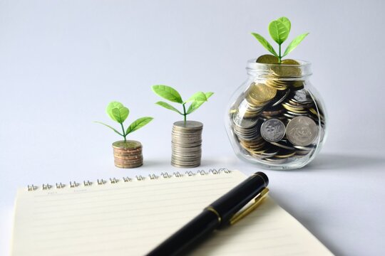 Eye-level close-up of dynamic coin stacks and a overflowing glass jar with small green plants placed behind a notebook and elegant pen, depicting banking growth and compound interest concepts.