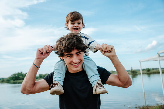 Man carries his baby brother on his shoulder near a summer river