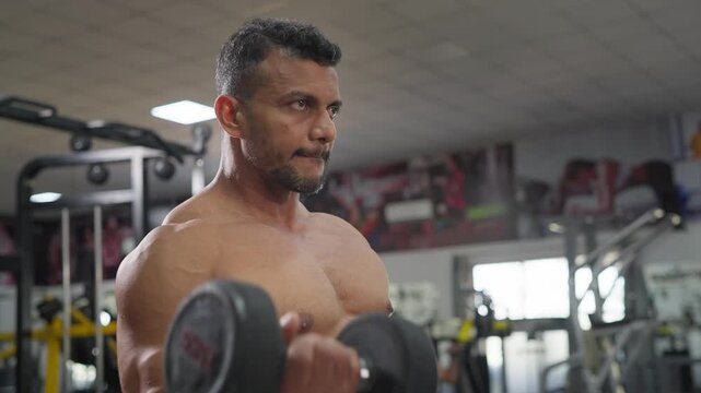 A middle aged Indian Asian fit muscular shirtless man performs a single arm dumbbell biceps curl exercise in a modern gym. Fitness, resistance workout, weight training, healthy lifestyle concept