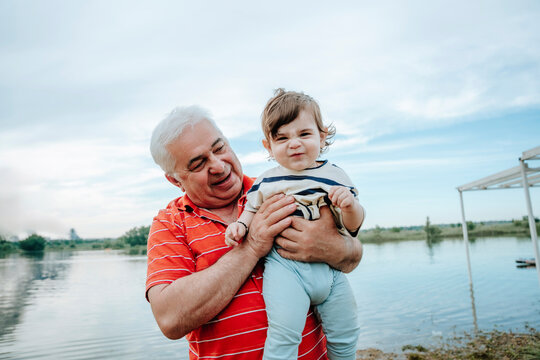 Smiling grandfather holds his grandson by a pond outdoors