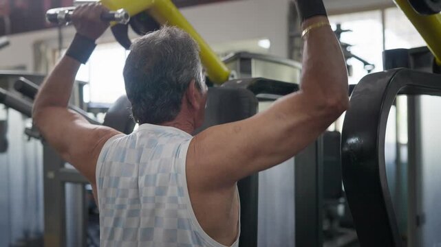 Indian Asian fit muscular elderly man using a lat pulldown machine to perform a resistance strength training exercise targeting the back and shoulders in modern gym. Fitness, workout, wellness concept