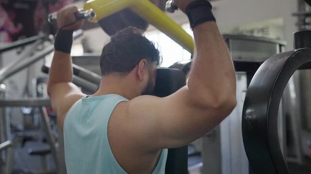 A young Indian Asian fit muscular man using a lat pulldown machine to perform a resistance weight training exercise targeting the back and shoulders in a modern gym. Fitness, workout, wellness concept
