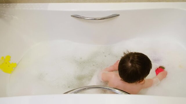 Toddler child playing with bubble in bathtub. Little boy sitting in soapy water. Hygiene routine and childcare at home. Water game, relaxation and clean concept for bath time.