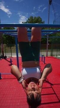 A fit woman performs a core workout, hanging from monkey bars at an outdoor gym. She demonstrates strength and dedication during her fitness training session under a bright sky.