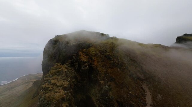 FPV drone glides down mist-covered volcanic cliffs in Iceland, revealing rugged basalt escarpments, moss-covered slopes, black coastal terrain and the North Atlantic during a fast descent.