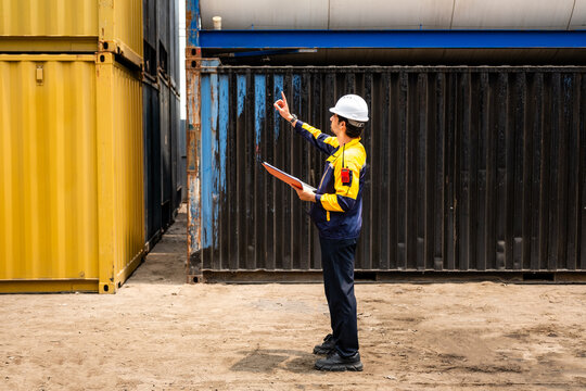 Safety-clad worker in a hard hat inspects and records stacked shipping containers in an industrial yard, holding a clipboard and using a handheld device.
