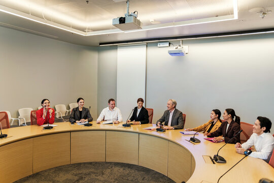 Group of professionals in a modern conference room holding a roundtable meeting with microphones and a projector screen, discussing plans and strategy.