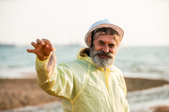 A bearded man in a hard hat and yellow raincoat smiles on the beach, proudly holding a small fish with the sea and shoreline in the background.