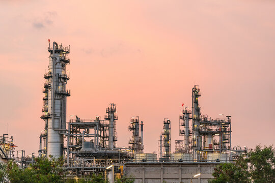 Petrochemical refinery with tall distillation towers, pipes, and platforms rising above greenery against a soft sunset sky.