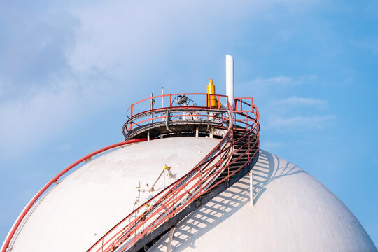 Close-up of a spherical industrial gas storage tank with red access stairs and safety railings against a blue sky.