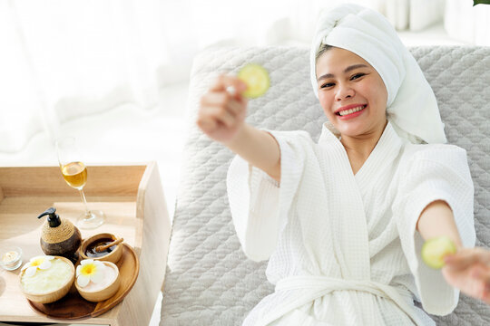 Smiling woman in a bathrobe and towel relaxes at a bright spa, holding cucumber slices, with a tray of skincare products, flowers, and a glass of champagne beside her.