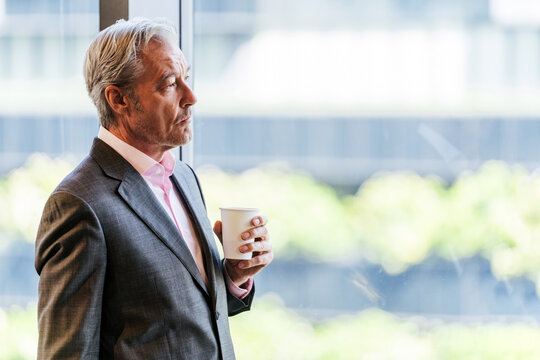 Mature business professional in a suit stands by a bright office window, holding a coffee cup and looking thoughtful during a break.