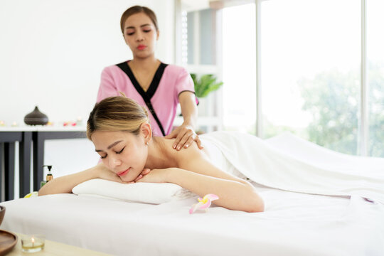 A woman relaxes during a professional back massage at a bright spa, while a therapist in pink uniform works gently in a calm, minimalist treatment room.